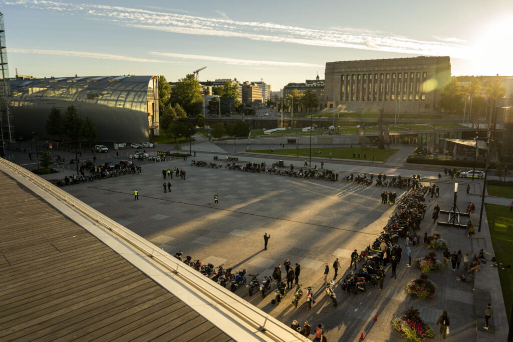 Kymmenittäin mopoja ja mopoilijoita mielenosoituksessa kansalaistorilla laskevan auringon valossa